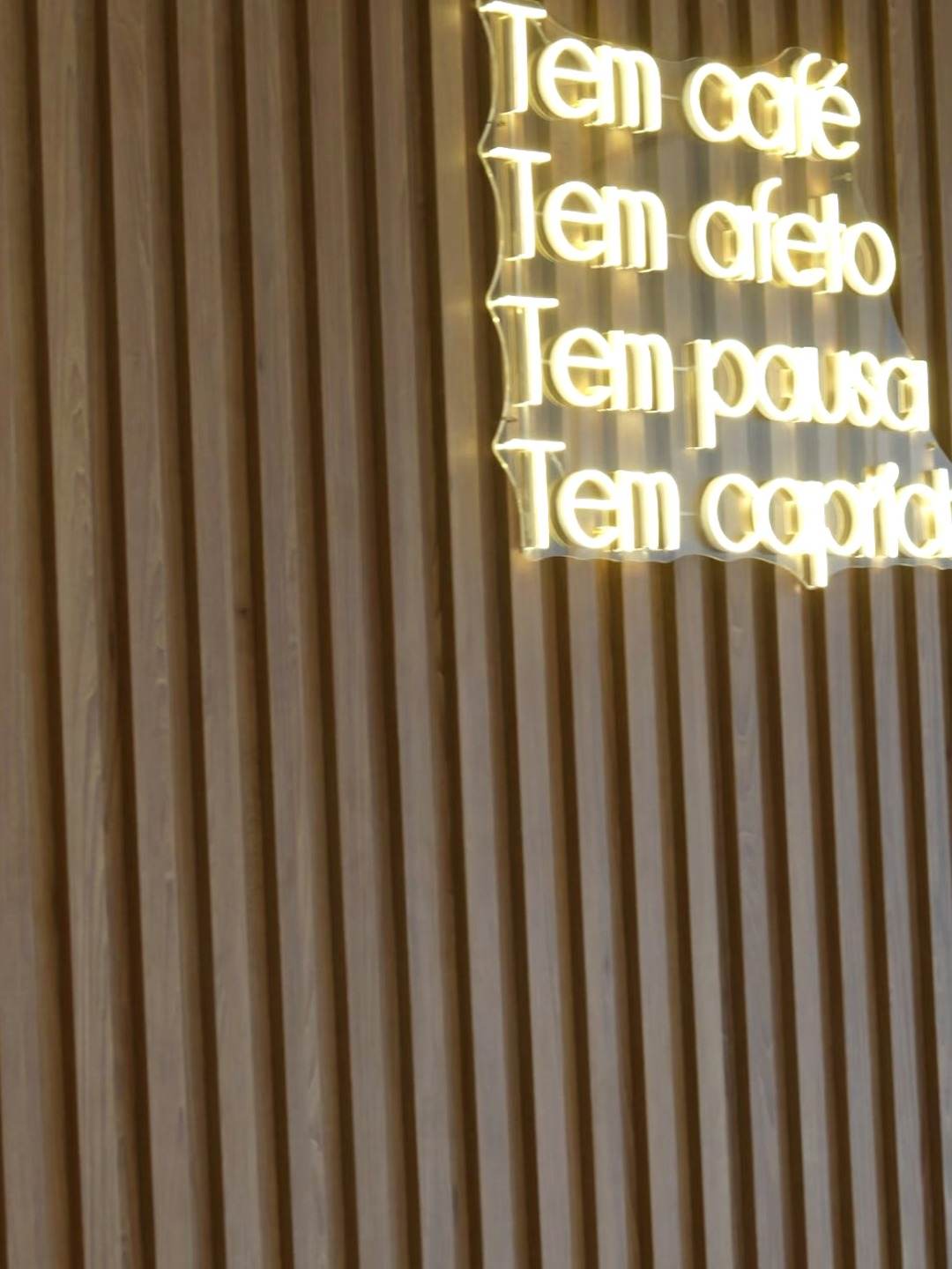 Painel ripado amadeirado com o neon "Tem café, tem afeto, tem pausa, tem capricho"