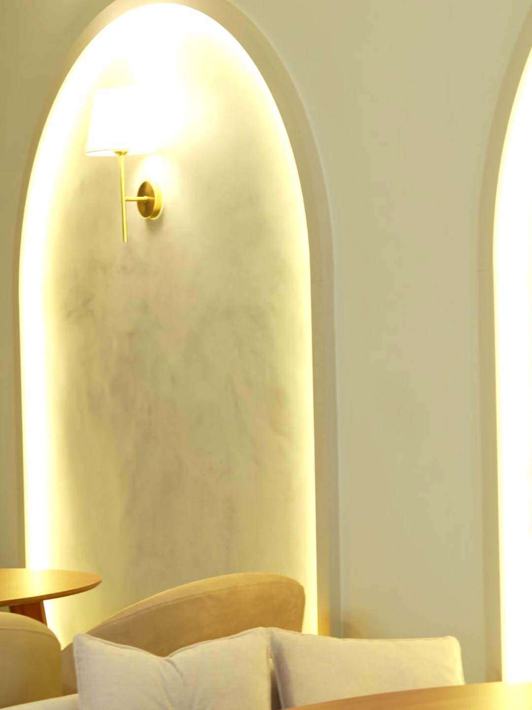 Arco iluminado na parede, com arandela dourada, sobre o espaço das mesas
